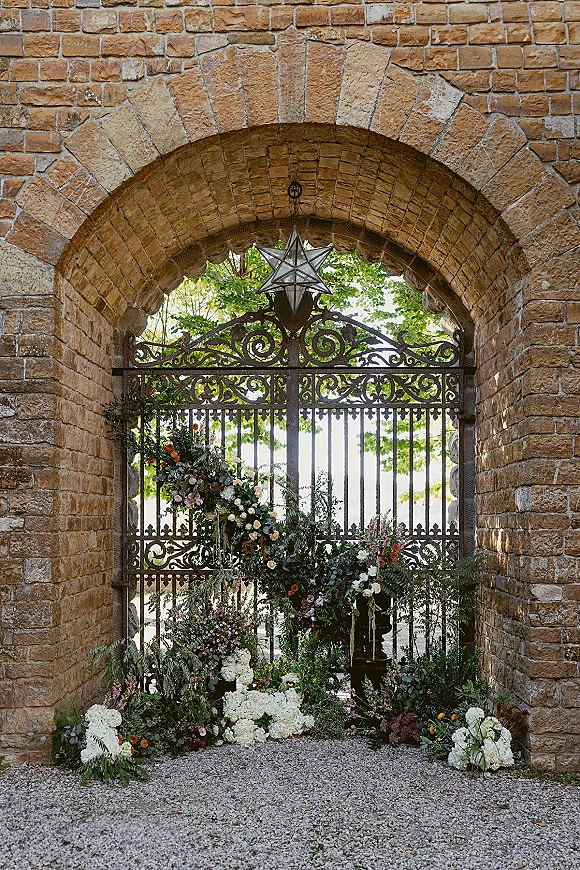 Wedding ceremony backdrop with wrought iron gate under a stone arch, hydrangea florals, greenery garland, and a hanging star lantern