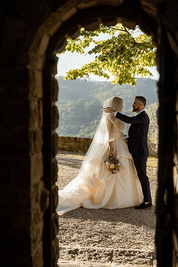 First look moment as groom lifts bride’s veil under a stone archway, her gown train and bouquet framed by mountains behind