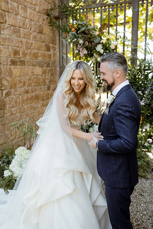 Couple portrait of bride and groom laughing, holding hands as her veil flows beside a sunlit stone wall and garden gate backdrop