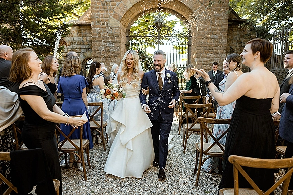 Wedding recessional as bride and groom walk the aisle through a wedding confetti toss, bouquet and veil flowing beneath a stone archway