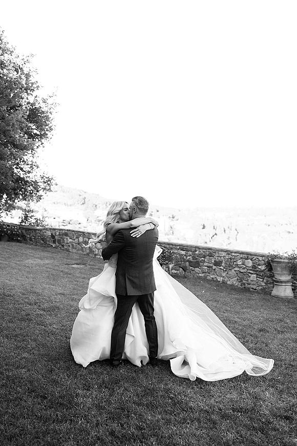 Wedding kiss portrait of bride and groom kissing as her long veil and dress train drape on a lawn by a stone wall and hillside view