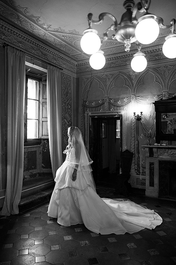 Bridal portrait in black and white of a bride by window, wearing a cathedral veil and long train in an ornate vintage room with chandelier