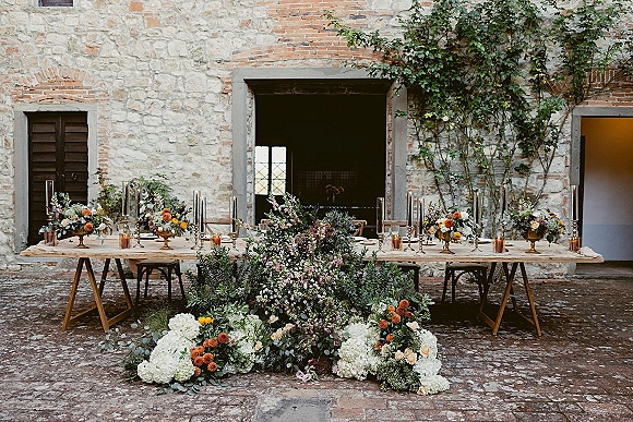 Reception tablescape with long banquet table decor, lush floral runner and tall taper candles in a cobblestone courtyard by a stone wall
