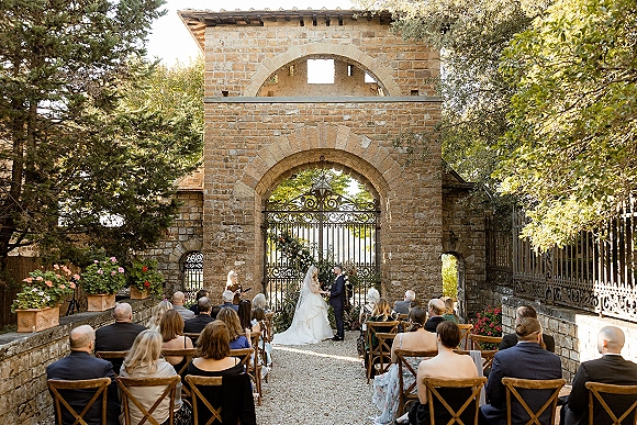 Wedding ceremony with bride and groom exchanging vows under a floral arch, long veil flowing in a stone courtyard with guests seated