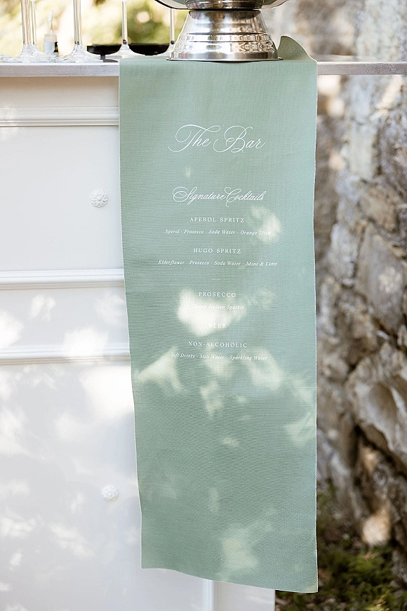 Bar menu sign with calligraphy wedding bar menu on a sage green runner beside champagne flutes and a silver dispenser at an outdoor bar counter