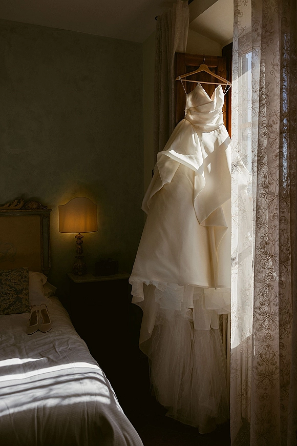 Wedding dress on hanger glowing in window light, tiered tulle skirt cascading above a bedroom bed with bridal shoes nearby