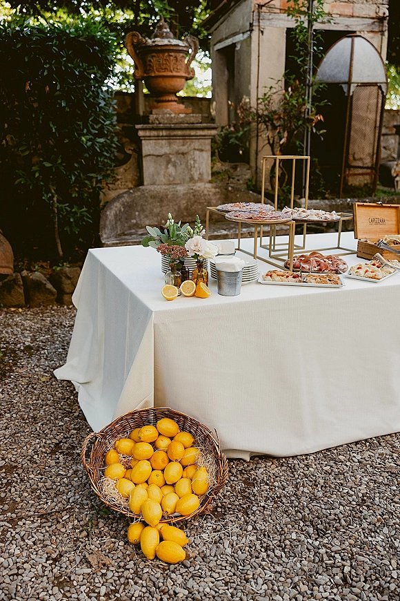 Wedding appetizer table with outdoor cocktail hour table styling, tiered stands of charcuterie and pizza, bud vases, and amber bottles in a garden courtyard