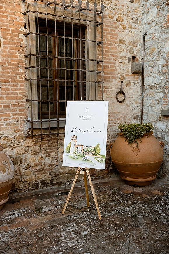 Wedding welcome sign on easel with custom illustrated venue artwork beside a terracotta urn planter and greenery in a stone courtyard backdrop