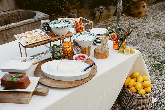 Wedding appetizer table with caprese skewer platter, olives, and citrus slices on wooden risers over white linen in a garden setting
