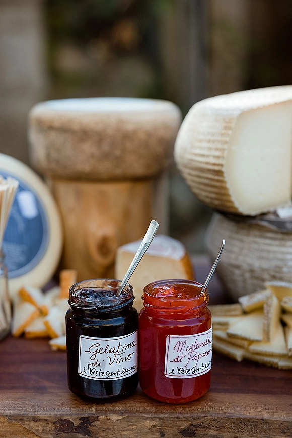 Cheese board with cheese wheels and wedges, crackers, and jam jars with spoons arranged on a wooden board in a blurred outdoor setting