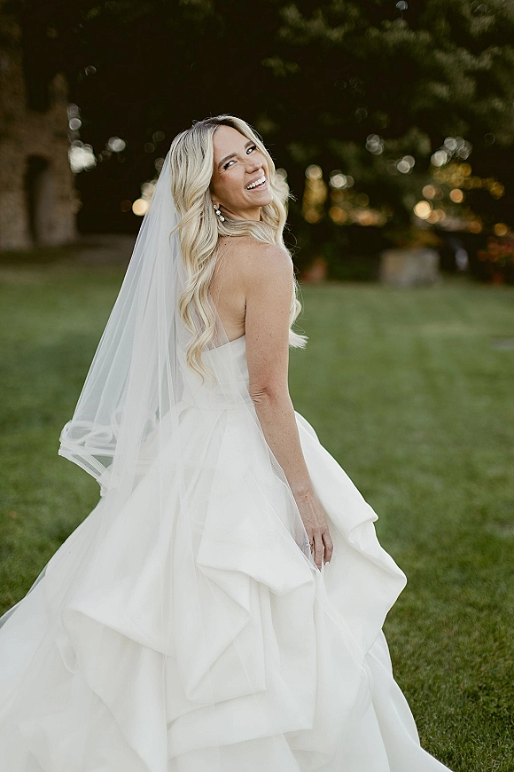 Bridal portrait of a smiling bride looking over her shoulder in a strapless wedding dress and veil, standing on a lawn by trees and a stone building