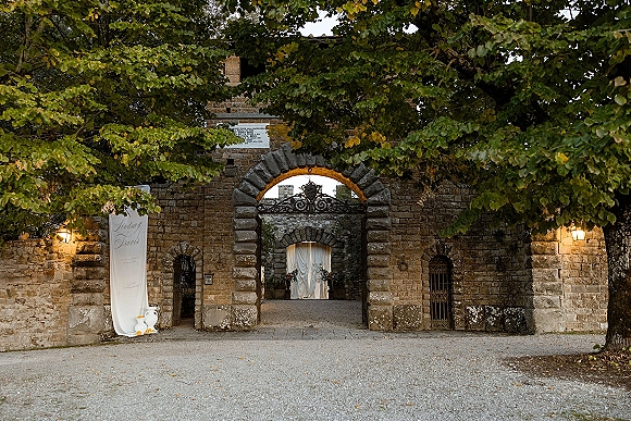 Wedding ceremony entrance framed by stone arch wedding entrance with draped fabric, floral urns, and a welcome banner in a gravel courtyard