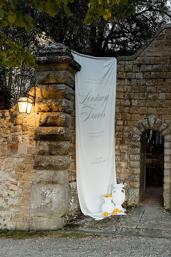 Wedding welcome sign on a draped fabric banner with calligraphy lettering, styled with white ceramic vases and citrus by a stone gate archway
