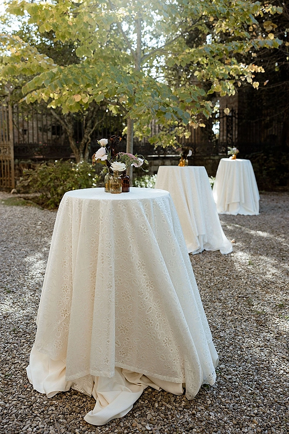 Cocktail table decor with white linens, lace tablecloth, and amber glass bud vases holding florals on a gravel garden patio backdrop