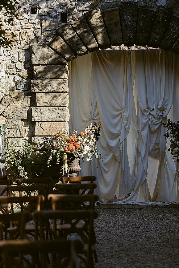 Ceremony backdrop with draped ceremony backdrop, floral urn pedestal and greenery framing a stone archway with wooden chairs in sunlight