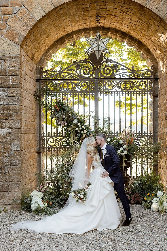Wedding kiss portrait of bride and groom kissing under a stone archway, her veil trailing as he holds her near florals and lanterns