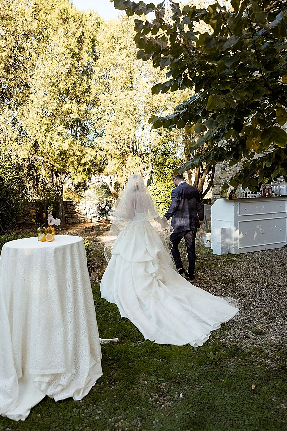 Couple walking portrait with bride and groom walking away, her long train and veil flowing down a sunlit gravel garden path by stone wall