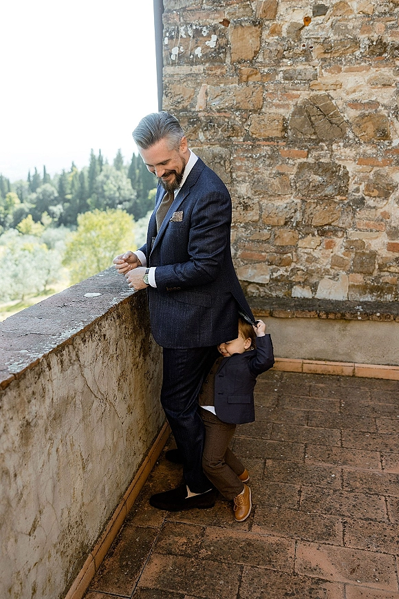 Groom portrait with child as a toddler hugs him in a navy suit on a terrace by a stone wall and railing, trees behind