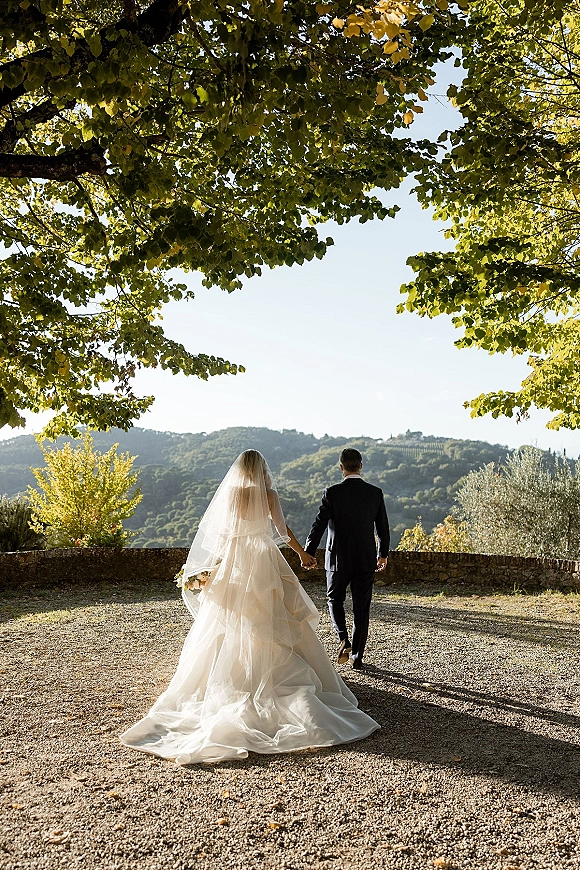 Couple portrait of bride and groom walking away, holding hands, bride’s veil and train flowing on a gravel path under trees