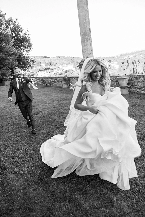 Wedding couple moment as the bride runs holding her dress while the groom chases behind on a lawn near a stone wall and column