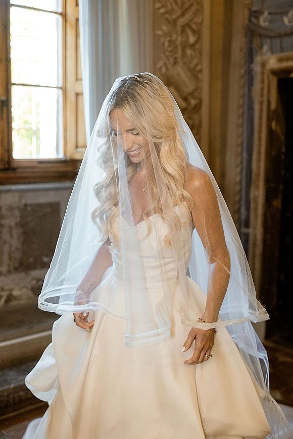 Bridal portrait of a bride wearing veil, looking down in a strapless gown with a cathedral veil, lit by a tall window in a formal room