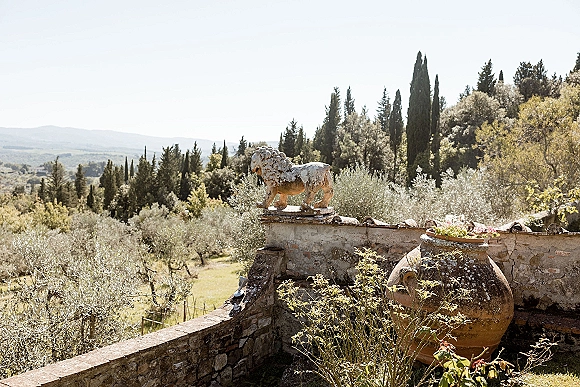 Wedding venue landscape with a stone wall, lion statue, and terracotta urn of flowers overlooking hills with cypress and olive trees