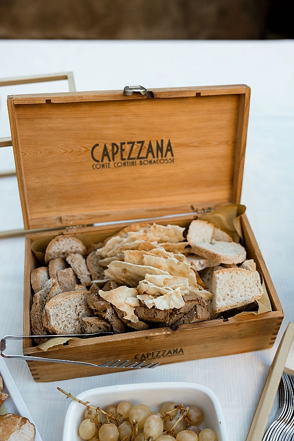 Wedding bread display with assorted sliced bread and flatbread crackers in a wooden box, with grapes and tongs on a white tablecloth