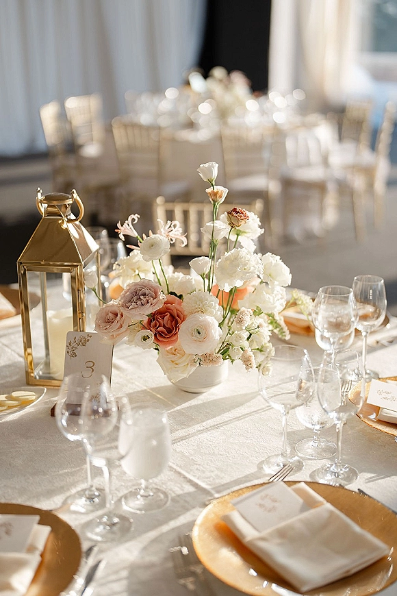 Reception tablescape with wedding table centerpiece, gold lantern, and white tablecloth with gold chargers, place cards, and glassware in window light
