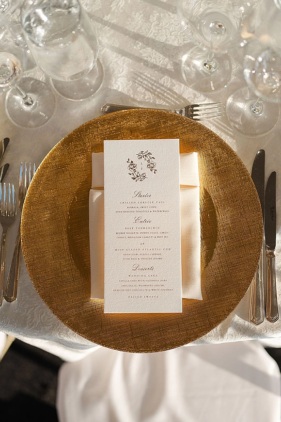Wedding place setting with a gold charger wedding menu card atop a folded white napkin, silver flatware and wine glasses on patterned linen tablecloth