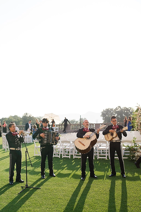 Wedding mariachi band playing trumpet and accordion in charro suits with red bow ties beside white ceremony chairs on a bright lawn