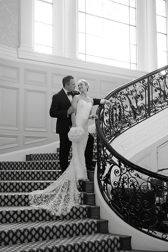 Couple portrait on a grand staircase, groom in black tux holding bride in strapless lace dress with long train by wrought iron railing