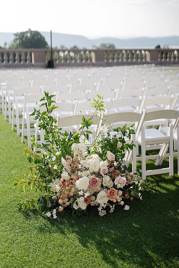 Ceremony aisle decor with roses and greenery in a ground floral arrangement beside white folding chairs on a lawn terrace with hills beyond