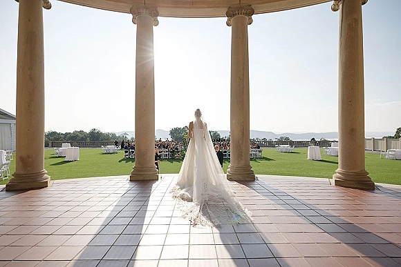 Bridal processional as bride walking down aisle in a wedding dress with cathedral veil and train, past chairs on a stone terrace with water view