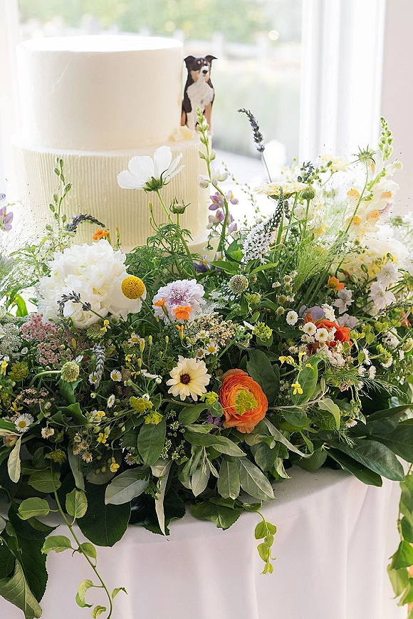 Wedding cake with textured buttercream, a two tier wedding cake topped with a figurine and wildflowers, lit by a window with greenery beyond