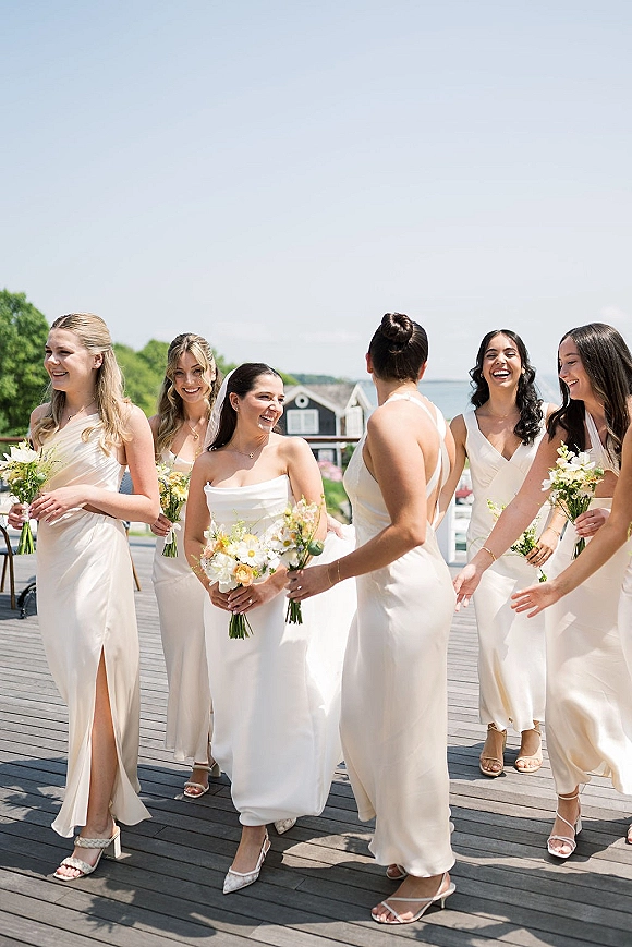 Bridesmaid group in champagne bridesmaid dresses walking and laughing with white and peach bouquets on a wood deck by the ocean