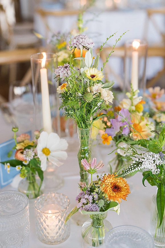 Wedding centerpieces with wildflower wedding centerpiece bud vases, greenery, and taper candles in glass hurricanes on softly lit reception tables
