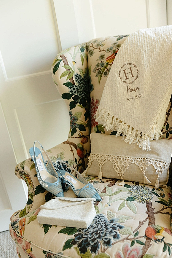 Bridal shoes in powder blue slingback heels beside an embroidered clutch on a floral armchair, set against a white paneled wall