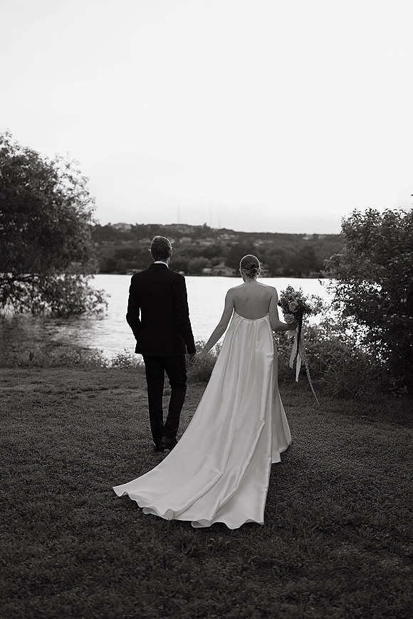 Couple portrait of bride and groom walking away hand in hand, her dress train and ribbon bouquet flowing by a lakeshore backdrop