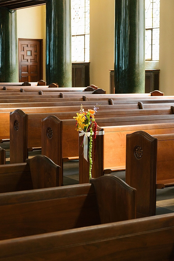 Church ceremony decor with wedding pew flowers tied in a ribbon bow, bright floral arrangement on wooden pews beneath stained glass windows