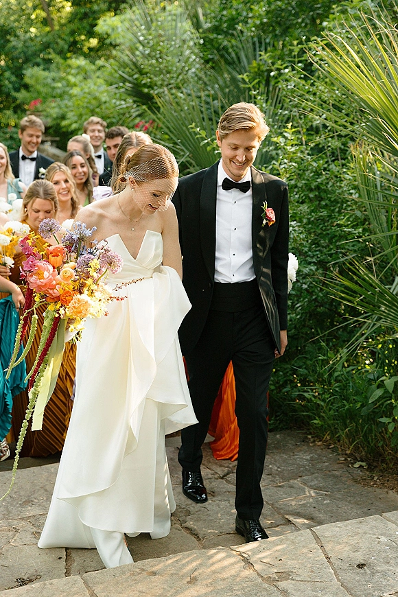 Wedding recessional as bride and groom walking down a stone garden path, she holds a colorful bouquet with ribbon streamers while guests cheer