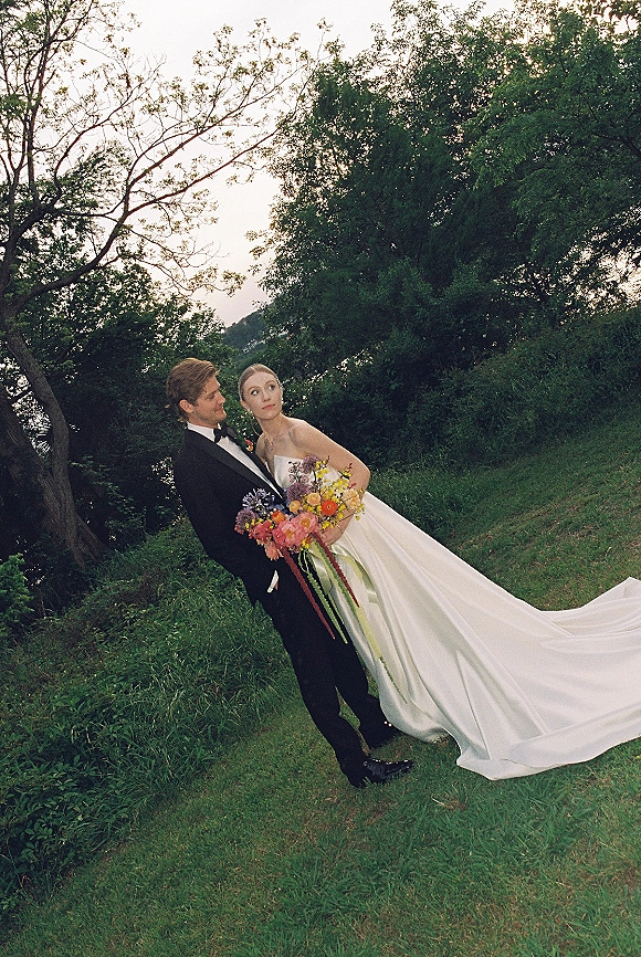 Couple portrait of bride in strapless gown with long train and colorful bouquet beside groom in tuxedo on grassy hill by lake