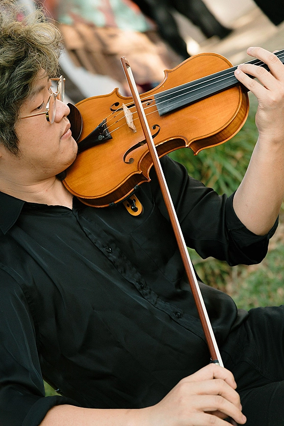 Wedding violinist in black shirt and eyeglasses playing violin with bow during an outdoor ceremony, greenery and blurred seating behind