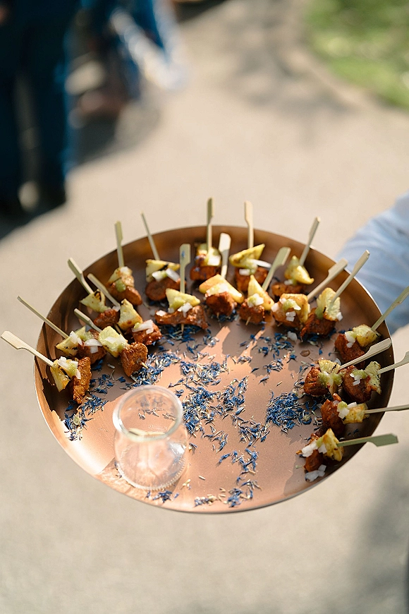 Wedding passed appetizers on a tray with cocktail skewers and savory bites, held by a caterer outdoors with greenery and guests blurred behind
