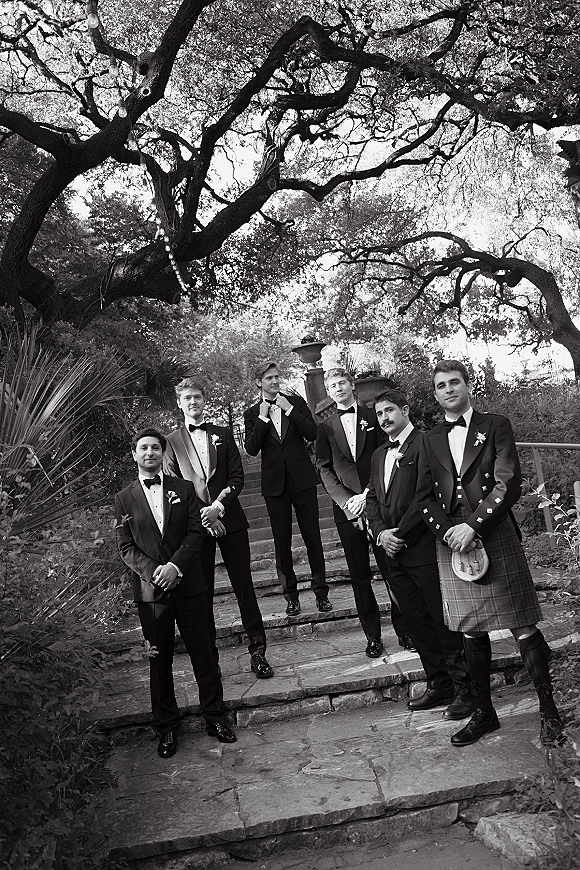 Groomsmen portrait of the groom with groomsmen in black tuxedos and one in a kilt on stone steps beneath oak and string lights