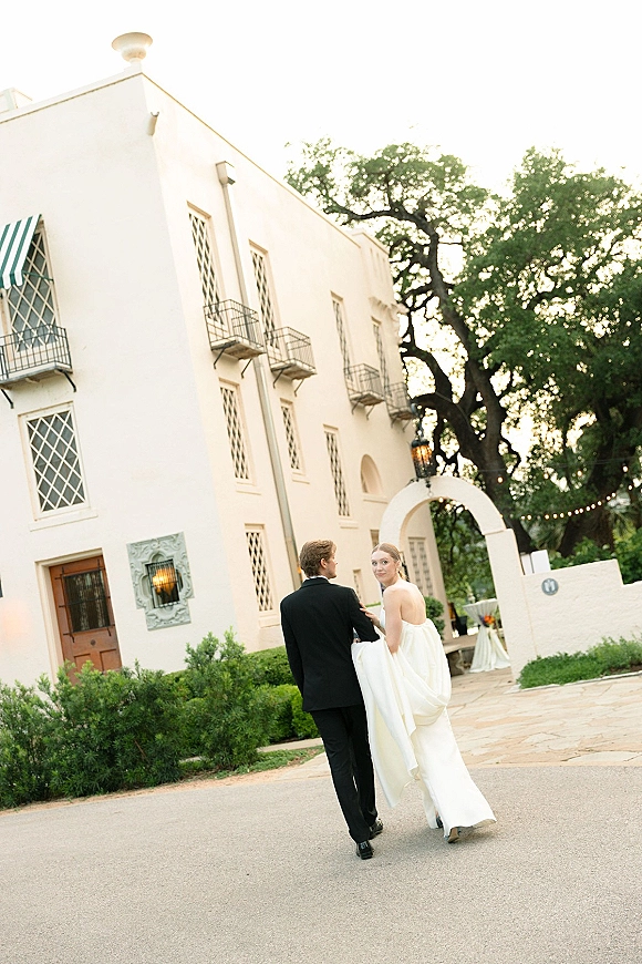 Couple portrait of bride and groom walking away as the bride looks back, holding her gown train in a stucco courtyard with archway