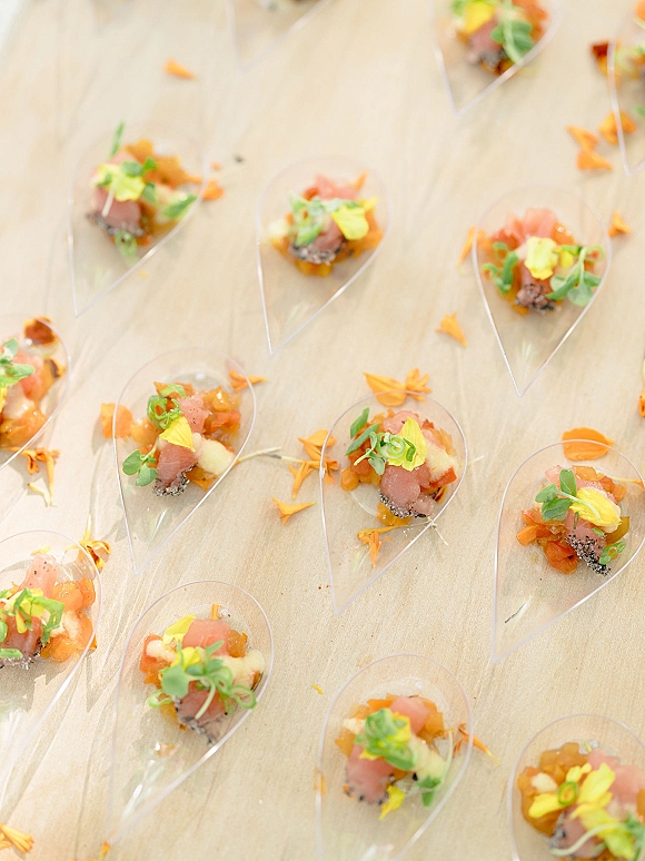 Wedding appetizers in clear plastic spoons with passed hors d'oeuvres, smoked fish, diced fruit, microgreens, and edible flowers on light wood surface