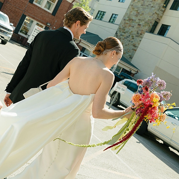 Wedding couple portrait of bride and groom walking away on a sunny city street, bride in birdcage veil holding a colorful bouquet