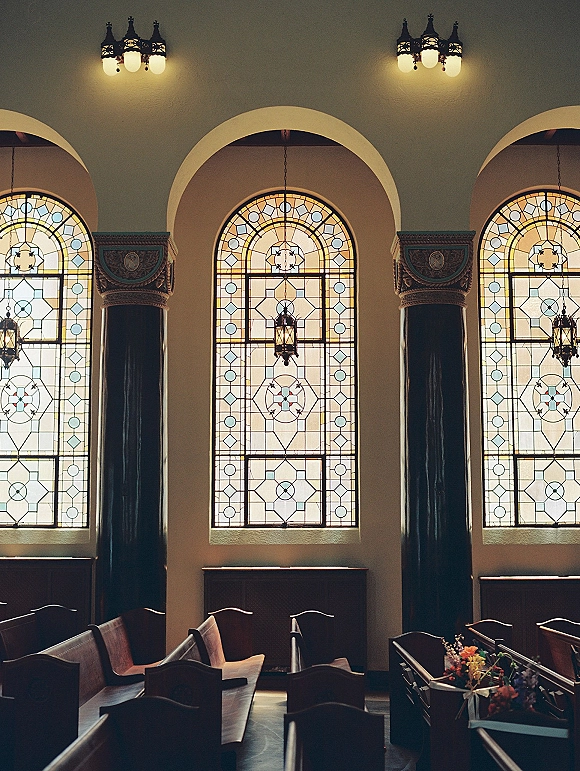 Ceremony venue interior with stained glass windows, wooden pews and floral pew markers, warm lantern lighting, and arched columns
