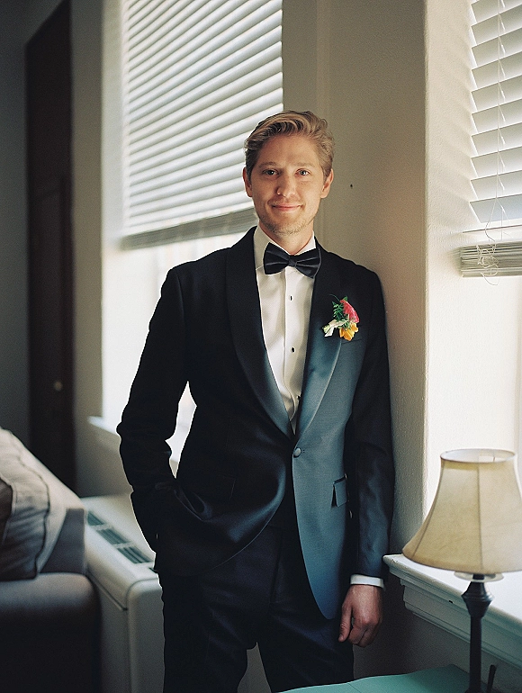 Groom portrait in a black tuxedo with bow tie and boutonniere, standing by window blinds in soft indoor light near a sofa and lamp