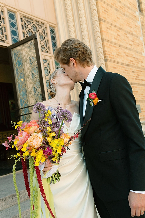 Wedding kiss portrait of bride and groom kiss, bride holding colorful bouquet and veil by an ornate doorway with brick and patterned windows